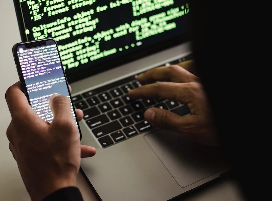 unrecognizable hacker with smartphone typing on laptop at desk